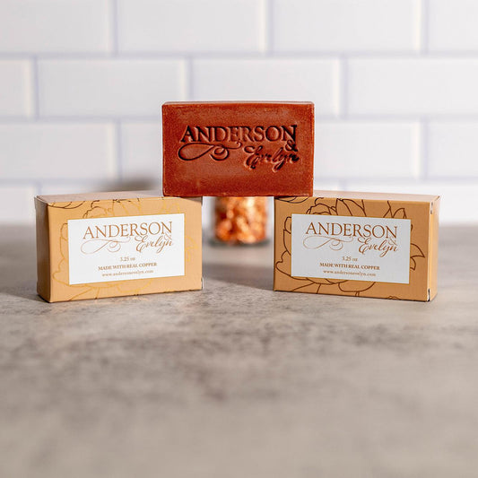 All-natural copper soap bar and packaging on marble counter with white tile background.
