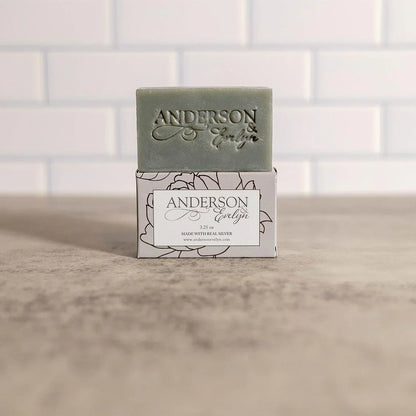 Natural green soap bar with engraved text, placed on floral box, white tile background.