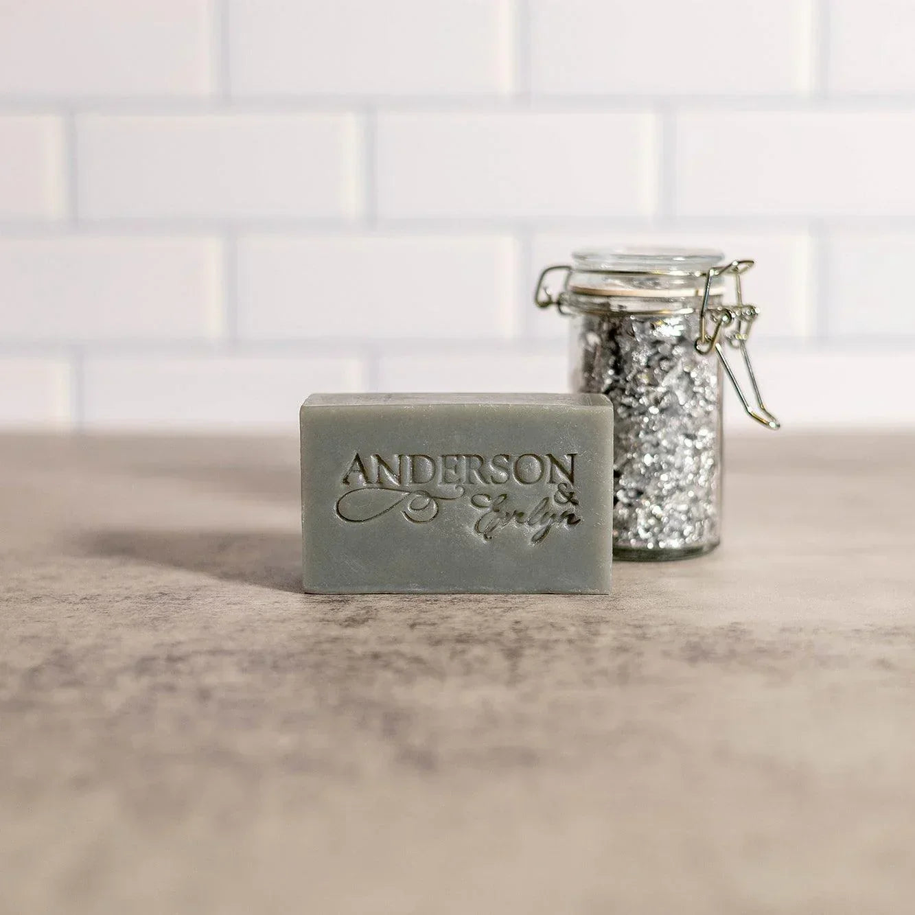 Gray natural soap bar with embossed text beside a glass jar of metallic flakes on countertop