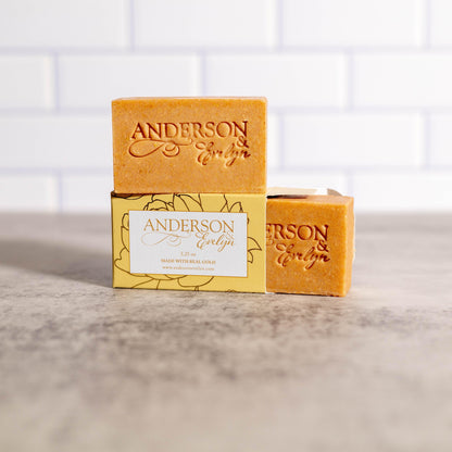 Natural bar soaps with gold hue and floral packaging on a marble countertop, kitchen tile background.