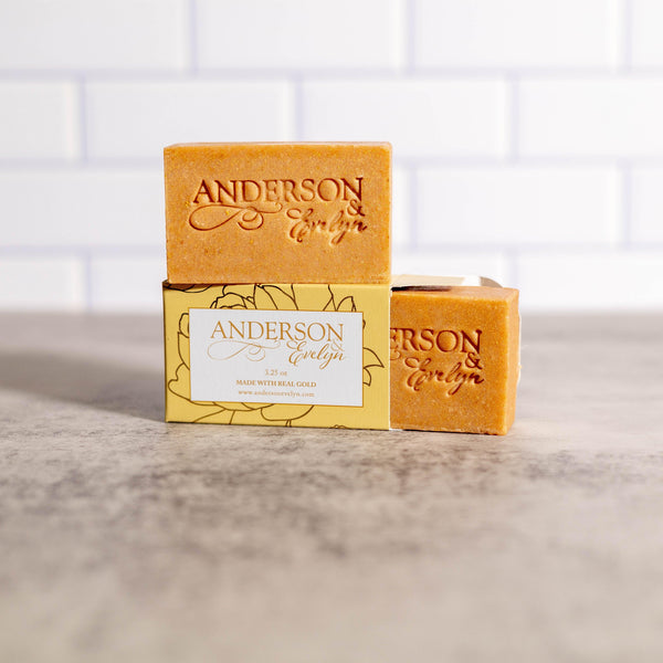 Natural bar soaps with gold hue and floral packaging on a marble countertop, kitchen tile background.