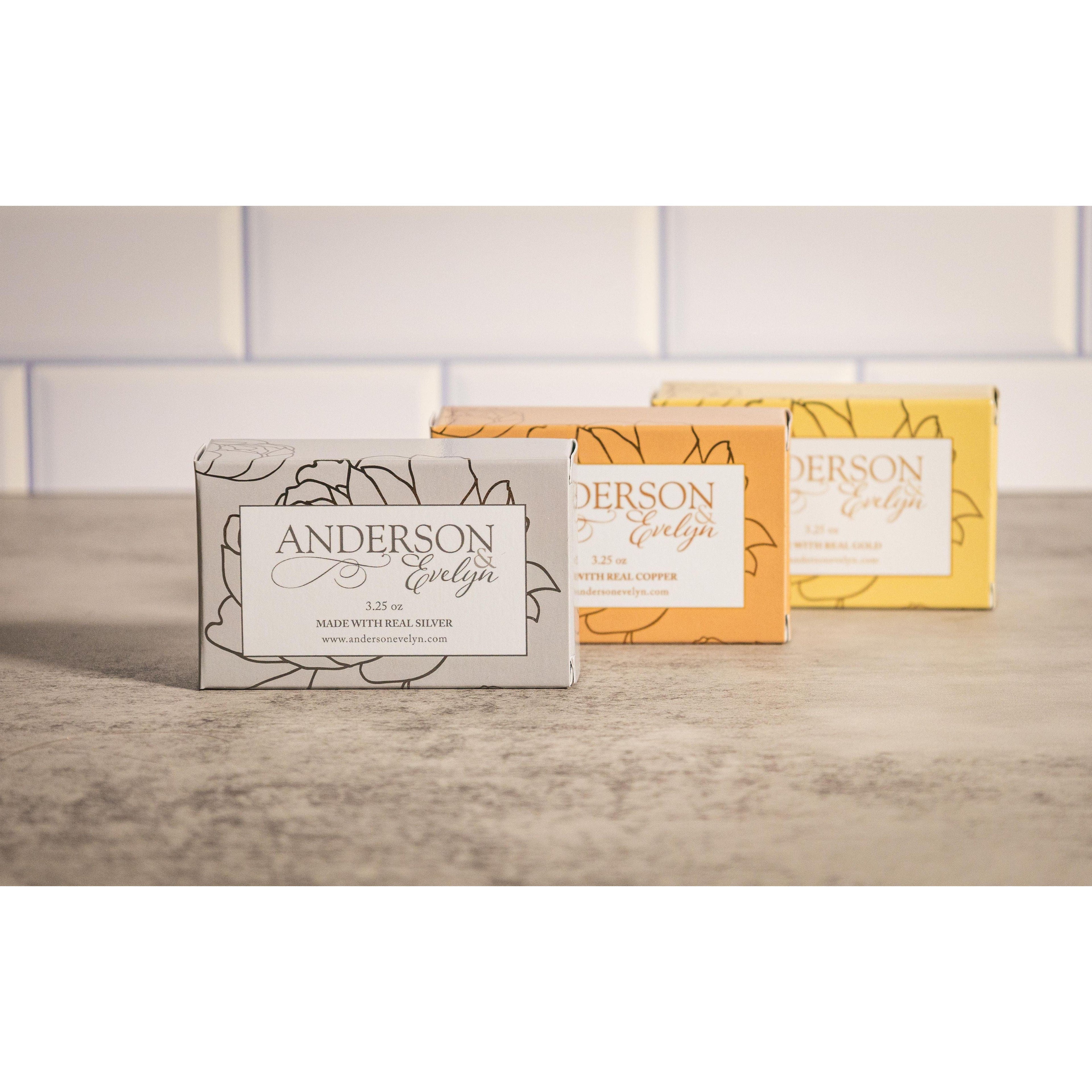 Three natural soap bars with silver, copper, and gold boxed packaging on a stone counter.
