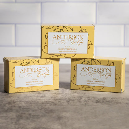 Three yellow boxes of all-natural soap with gold accents stacked on a marble surface