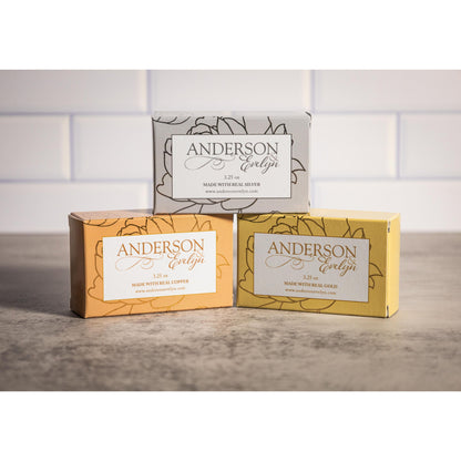 Three boxed natural soaps infused with copper, silver, and gold ions on a stone surface.