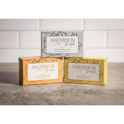 Three bars of all-natural soap with copper, silver, and gold ion-infused packaging on a countertop.